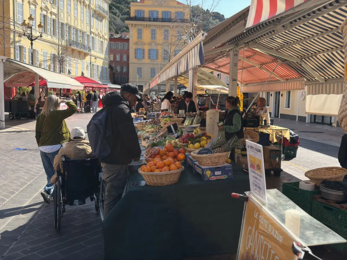 Des promeneurs devant les étals de fruits et légumes du cours Saleya, façades ocre en arrière-plan.