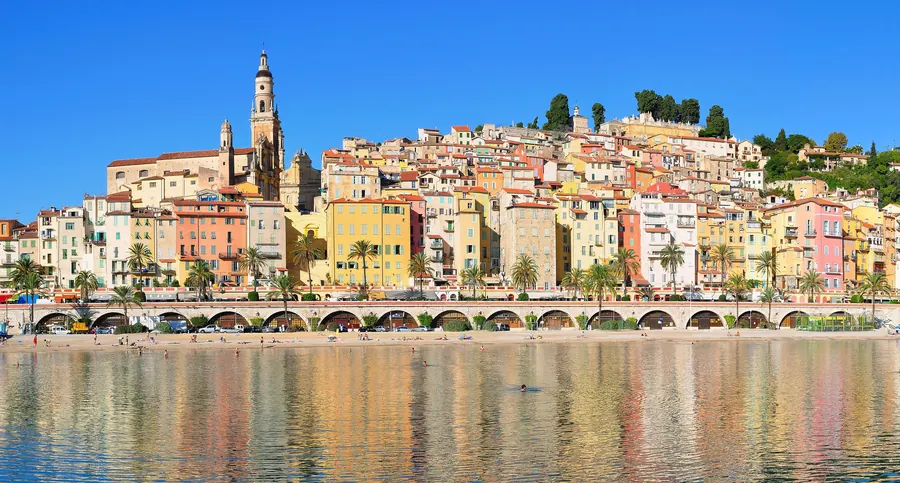 La vieille ville de Menton, façades jaunes, ocre et roses dominées par le clocher de la basilique, reflétées dans la mer.
