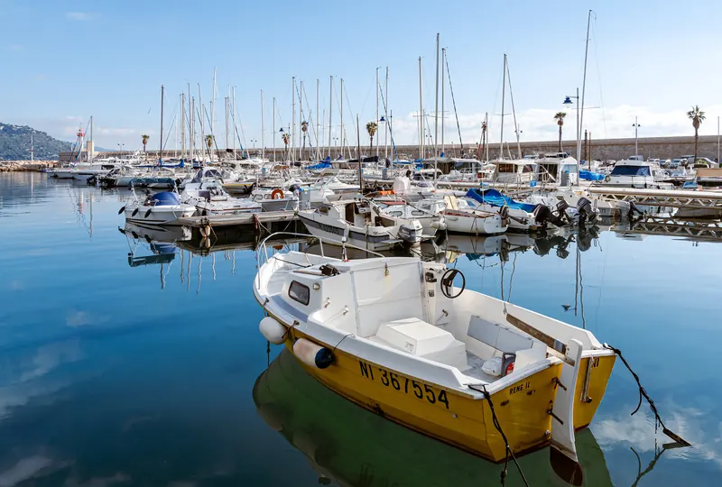 Une barque jaune amarrée sur l'eau immobile du port de Menton, voiliers en arrière-plan.