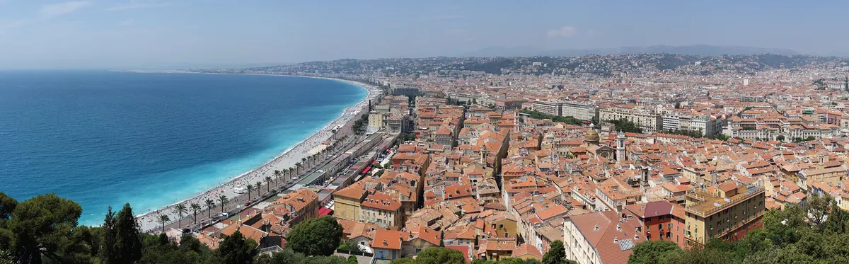 La baie des Anges à Nice vue depuis la colline du Château : la mer turquoise, la plage de galets et les toits de tuiles de la ville.