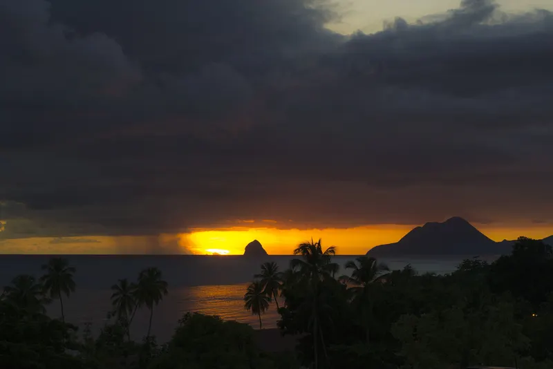 Le soleil se couche derrière le rocher du Diamant, sous un ciel d'orage, avec des cocotiers en ombre chinoise.